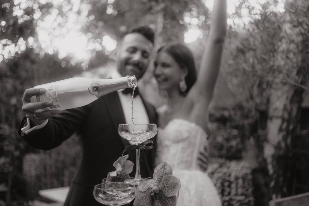 Fröhliches Brautpaar beim Anstoßen: Der Bräutigam gießt Sekt in Gläser, während die Braut jubelnd neben ihm steht – ein Moment voller Leichtigkeit und Glück.