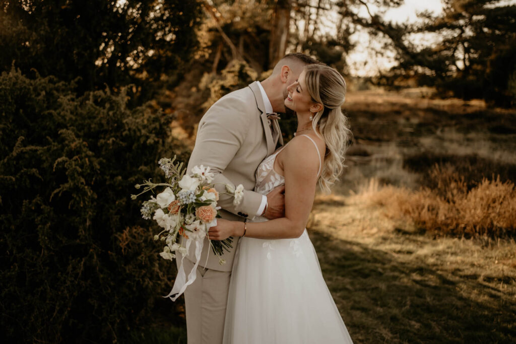 Braeutigam küsst die Braut liebevoll am Hals während eines natürlichen Paarshootings im Grünen, aufgenommen von einem Hochzeitsfotograf