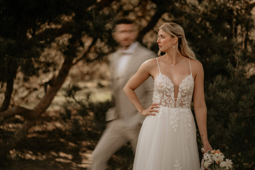 Braut im eleganten Spitzenkleid steht selbstbewusst im Fokus, während der Bräutigam unscharf im Hintergrund vorbeigeht – kreativ inszeniert von Hochzeitsfotograf Christian Messner.