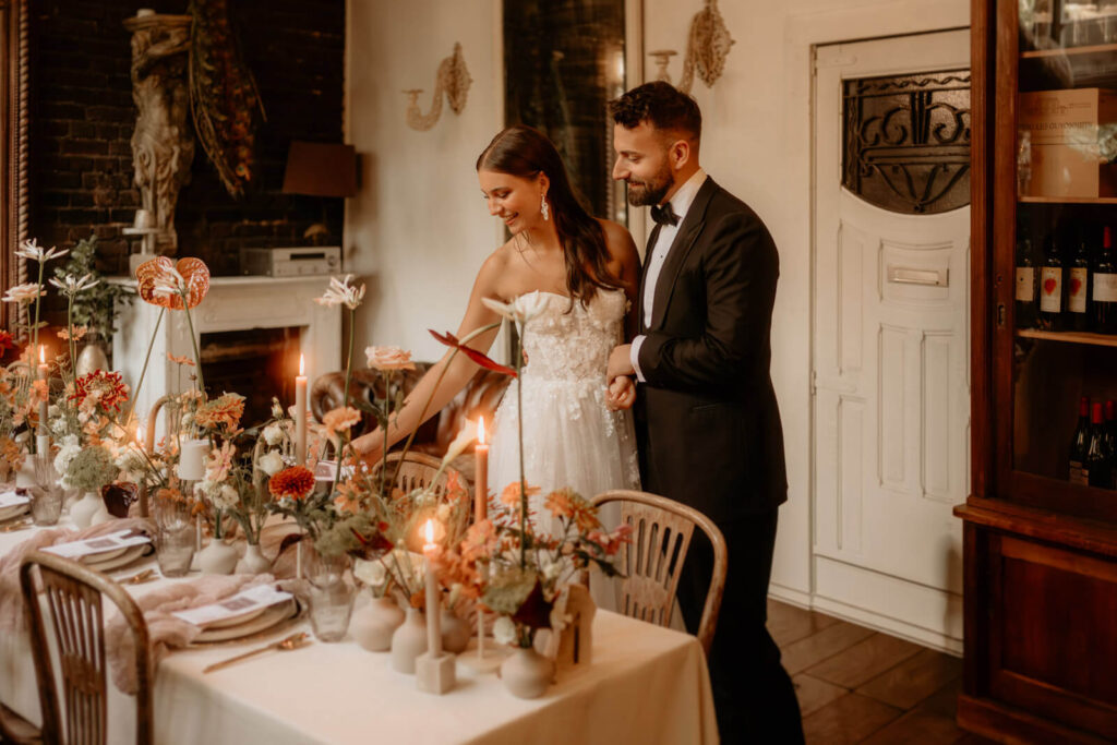 Romantischer Moment zwischen Braut und Bräutigam, die Hand in Hand die elegant gedeckte Tafel mit Kerzen und floralen Details betrachten.