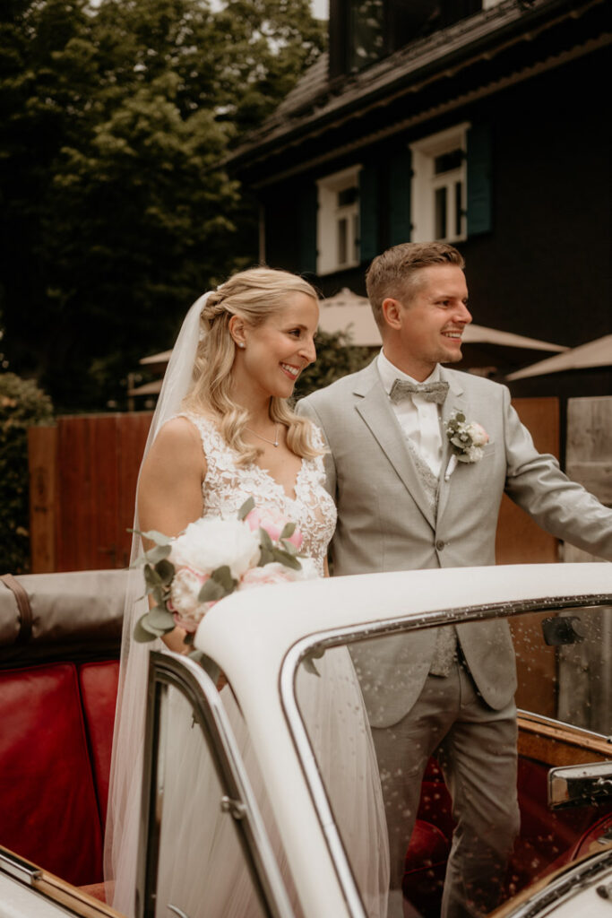 Glückliches Brautpaar steht gemeinsam am Oldtimer und genießt den besonderen Moment