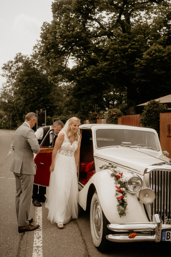 Christian Messner fotografiert das Brautpaar beim eleganten Ausstieg aus einem weißen Hochzeitsauto vor der Säge in Kappel