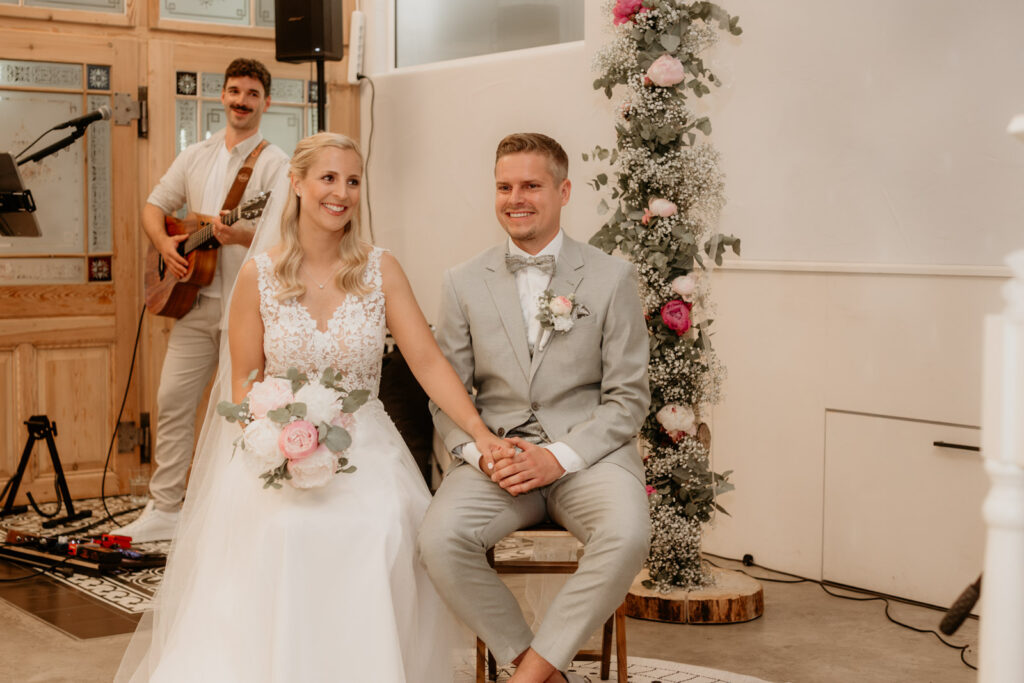 Braut und Bräutigam sitzen während der Trauung in der Säge in Kappel Hand in Hand, begleitet von Live-Musik und Blumenarrangements