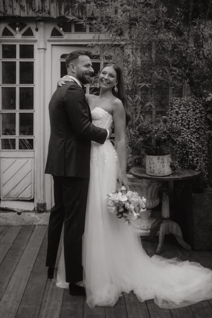 Fröhliches Brautpaar steht lachend vor einem alten Gartenhaus, umarmt sich zärtlich und genießt den Moment voller Liebe.