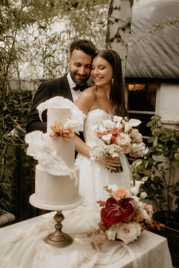 Christian Messner fotografiert ein glückliches Brautpaar, das liebevoll vor einer stilvollen Hochzeitstorte mit Blumenarrangement posiert.